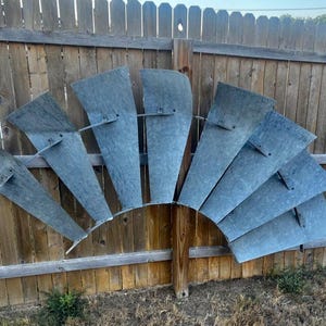 May include: A weathered, vintage windmill fan with eight galvanized metal blades. The blades are attached to a wooden post and mounted on a wooden fence. The blades are a faded gray color.