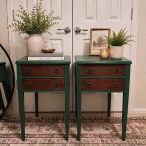 May include: A pair of matching forest green bedside tables, each featuring two drawers with dark wood fronts and brass-toned ring pulls. The tables stand on four straight legs and are topped with decorative items including vases, books, and a framed picture.