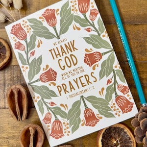 May include: A greeting card with the text "THANK GOD" and "PRAYERS" surrounded by floral illustrations in orange and green. A teal pencil rests on the wooden surface next to the card. Dried botanicals and a pinecone are also visible.