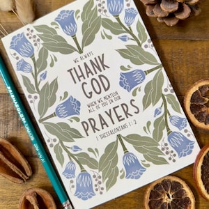 May include: A notebook with a floral design in blue and green, featuring the text "THANK GOD" and a Bible verse. A turquoise pencil and dried botanicals surround the notebook on a wooden surface.