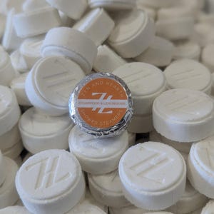 May include: A collection of white, circular shower steamers, each embossed with a "Z" and "H" logo. One is wrapped in silver foil, displaying "ZEN AND HEARTH" and "CEDARWOOD & LEMONGRASS". These are bath and body products, designed for use in the shower.