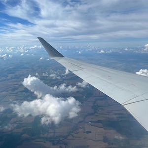 Könnte beinhalten: Ansicht aus einem Flugzeugfenster, die einen Flügel vor dem Hintergrund eines blauen Himmels mit Wolken zeigt. Die Landschaft darunter zeigt ein Flickwerk aus Feldern und Wäldern.
