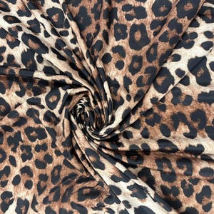 Pode incluir: Close-up de um tecido com estampa de leopardo. O tecido apresenta um fundo bege com manchas marrons escuras e pretas. O tecido é dobrado e drapeado, criando um efeito de redemoinho.