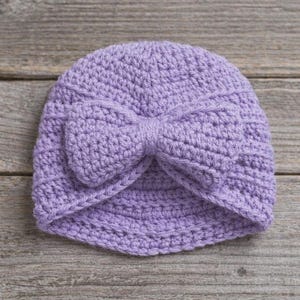 May include: A lavender crochet hat with a large bow detail. The handmade hat is made of soft yarn and is suitable for babies or children. The hat is shown on a rustic wooden surface.