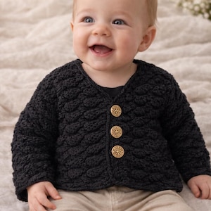 Op de afbeelding: Een lachende baby draagt een zwart, getextureerd gebreid vest met drie goudkleurige knopen. De baby draagt ook een beige broek. Het vest heeft een V-hals en lange mouwen. De achtergrond is een zachte, neutrale kleur.