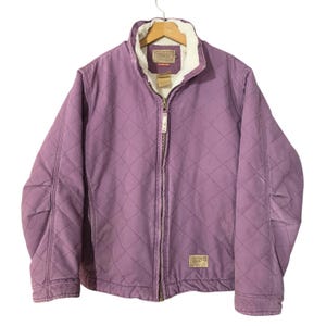 May include: A lavender quilted jacket featuring a full-length front zipper, a white sherpa fleece lining, and a stand-up collar. The exterior has a diamond-stitched pattern, long sleeves, and a rectangular brand patch on the lower front.