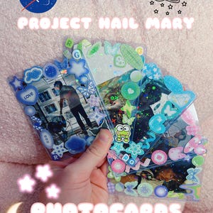 May include: Three rectangular clear plastic sleeves containing photographs, decorated with various holographic stickers including blue and green stars, hearts, butterflies, and circles. The text NASA and PROJECT HAIL MARY PHOTOCARDS is visible.