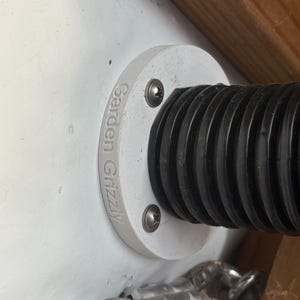 May include: A close-up of a white circular fitting with the engraved text "Garden Grizzly". The fitting is secured with two screws and is attached to a black, ribbed hose. The background is a white surface and wood.