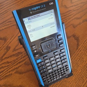 May include: A black and blue TI-nspire CX II calculator with a screen showing numerical data. The calculator is in a black stand on a wooden surface. The calculator has a full keyboard and the text "TEXAS INSTRUMENTS" is visible.