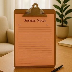 May include: A clipboard with a peach-colored paper, featuring the text "Session Notes" at the top. The paper has horizontal lines for writing. A black pen rests on the wooden table. The clipboard has a metal clip at the top.