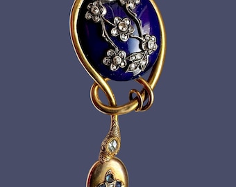 May include: An antique gold-toned brooch with a deep blue oval center. The center features a floral design with white gemstones. A smaller gold pendant with blue and white gemstones hangs below the oval.