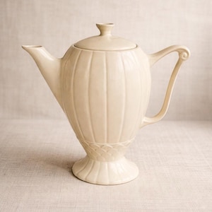 May include: Cream-colored ceramic teapot with a lid and a curved handle. The teapot has vertical ribbed detailing and sits on a pedestal base with a decorative pattern. The spout is angled, and the lid has a small knob.