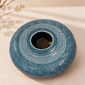 May include: A round, teal ceramic flower frog vase featuring a textured, repeating wave-like pattern on its surface. The center opening reveals a brass kenzan pin holder with sharp, upright metal needles designed for arranging stems securely.