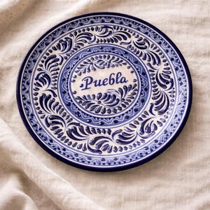 May include: A round, white ceramic plate with a blue floral design and the word "Puebla" in the center. The plate has a dark blue rim and is decorated with intricate patterns. The plate is likely handmade and is a decorative item.