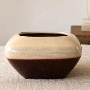 May include: A two-toned ceramic vase with a rectangular opening. The top half is a creamy beige, and the bottom half is a deep brown. The vase has a smooth, glossy finish and a rounded, geometric shape. This decorative vase is suitable for home decor.