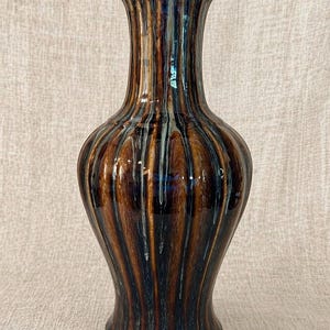 May include: A tall, decorative vase with a flared rim and a bulbous body. The vase features vertical ribbing and a glossy finish with a blend of brown, blue, and white tones. This ceramic vase is a stylish home decor item.
