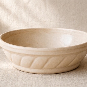May include: A beige ceramic mixing bowl with a decorative, ridged pattern around the exterior. The bowl is round and sits on a neutral-colored surface. The bowl is likely used for food preparation.