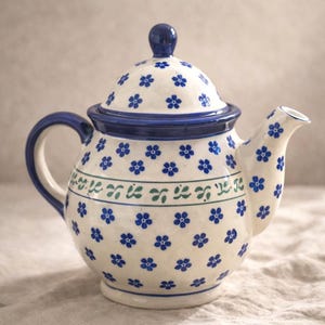 May include: A ceramic teapot with a white base, adorned with blue floral patterns. The teapot features a blue handle, rim, and lid, with a matching blue knob. A green and blue decorative band encircles the body.