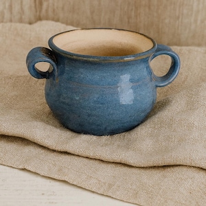 May include: A handmade, blue ceramic bowl with two handles. The bowl has a rounded shape and a light beige interior. The exterior is a matte blue with subtle variations in color. The bowl is sitting on a beige linen cloth.