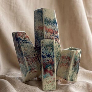 May include: A set of four ceramic vases with a speckled design in blue, red, and green on a cream background. The vases have geometric shapes, including rectangular and angled forms, and are arranged together.