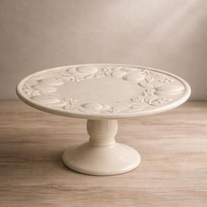 May include: Off-white ceramic cake stand with a round, raised platform and a pedestal base. The platform features a detailed embossed design of fruits and leaves. The stand is set on a light-colored wooden surface.