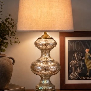 May include: A table lamp with a textured, metallic mercury glass base in a double-gourd shape, featuring a brass-toned metal base and finial, topped with a neutral-colored, cylindrical fabric lampshade that emits a warm, soft glow.