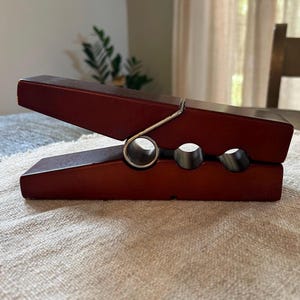 May include: A large, decorative wooden clothespin in a rich burgundy color. The clothespin features a silver-colored spring and two circular cutouts. It is resting on a textured, light-colored surface.