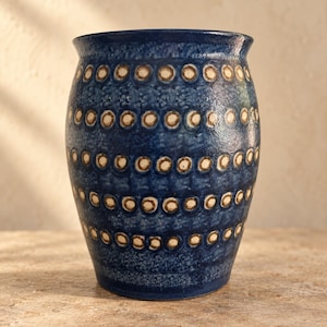 May include: A cylindrical ceramic vase with a deep blue glazed finish. The exterior features horizontal bands of repeating, recessed circular indentations, each containing a light beige center, creating a textured pattern around the vessel.
