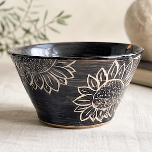 May include: A black ceramic bowl featuring a hand-carved sunflower pattern with light-colored outlines. The bowl has a wide, flared rim and a smooth, dark finish that contrasts with the natural clay color visible at the base and the carved floral details.