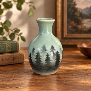 May include: A small ceramic vase with a smooth, light mint green glaze. The base of the vase features a dark, charcoal-colored gradient that transitions into a silhouette pattern of evergreen pine trees encircling the lower portion of the body.