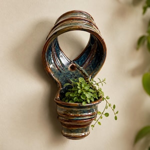 May include: A wall-mounted ceramic planter featuring a glossy, multi-toned glaze in shades of deep blue, brown, and copper. The vessel has a circular upper opening and a smaller, tapered lower basin containing a small, green leafy plant.