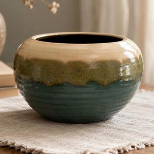 May include: A round, ceramic bowl with a dark interior and a three-tone exterior. The top is tan, transitioning to olive green, and then to a deep teal at the base. The bowl has a glossy finish and sits on a neutral-colored surface.