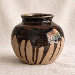 May include: A handcrafted ceramic vase with a rounded body and a dark brown, glossy glaze that drips down over a tan base. The vase has a small opening at the top and is likely used for decorative purposes.