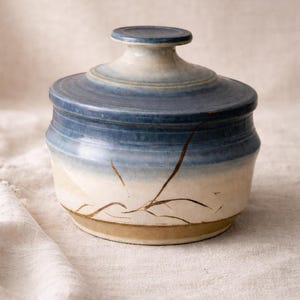 May include: A ceramic jar with a lid, featuring a gradient of blue and white hues. The jar has a round shape with a decorative brown line design. The lid has a small handle on top. The jar is sitting on a white cloth.