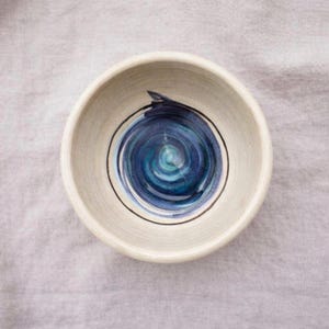 May include: A small, off-white ceramic bowl with a hand-painted blue spiral design in the center. The bowl has a simple, rounded shape and a black outline around the spiral. The bowl is sitting on a light gray surface.