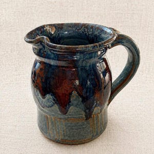 May include: A handmade ceramic pitcher with a dark blue and brown glaze. The pitcher has a rounded body, a spout, and a curved handle. The glaze has a glossy finish, with drips of color adding texture.