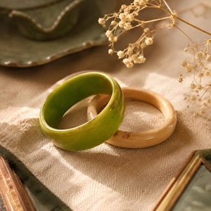 May include: Two circular bangle bracelets are displayed. One is a thick, polished, solid green band with subtle tonal variations. The second is a thinner, smooth, cream-colored band with light brown marbling throughout its rounded surface.