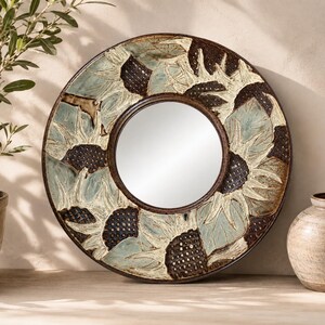 May include: A round wall mirror featuring a wide, decorative frame with a relief pattern of sunflowers. The frame displays a color palette of muted sage green, cream, and dark brown, with textured centers on the flowers and smooth, carved petals.