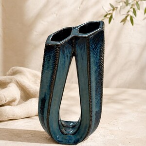 May include: A tall, dark blue ceramic vase with a unique U-shaped, open-center design. The surface features a glossy, mottled glaze with deep indigo tones and textured, dark brown edges along the vertical seams, creating a distinct geometric silhouette.