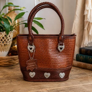 May include: A brown leather handbag with a crocodile-embossed texture, featuring braided handles, silver-tone heart-shaped hardware, and a tag labeled Genuine Leather. The base is accented with three silver heart studs in a darker brown leather trim.