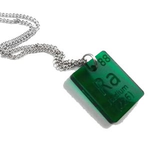 May include: A silver-toned metal chain necklace featuring a rectangular, translucent green pendant. The pendant displays the chemical element symbol Ra, the atomic number 88, the name Radium, and the atomic mass 226 in a dark green font.