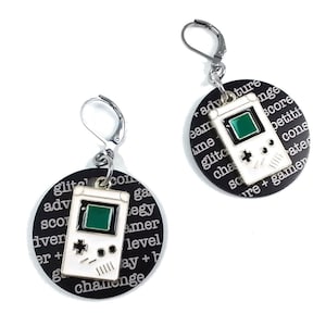 May include: A pair of black and white earrings with a silver hook. Each earring features a small white Game Boy with a green screen and black buttons. The earrings are surrounded by a black circle with white text that reads "adventure, score, gamer, level, play + challenge, glitch, console, team, time, score, petition, character, coins, rate, score + gamer".