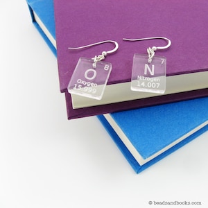 May include: A pair of silver dangle earrings with clear acrylic charms featuring the chemical element symbols for oxygen (O) and nitrogen (N). The charms are engraved with the element's name, atomic number, and atomic weight.