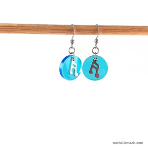 May include: A pair of dangle earrings featuring translucent bright blue circular acrylic discs. Each disc is accented with a silver-toned metal musical note charm suspended from a silver-toned hook wire. The design is simple and symmetrical.