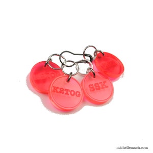 May include: Four bright pink plastic stitch markers with metal rings. The markers are labelled "k2tog", "ssk", "sl1, k1, psso", and "p2tog".