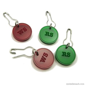 May include: Four round, plastic stitch markers with safety pins. Two markers are red with "WS" printed on them and two are green with "RS" printed on them.