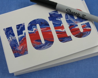 May include: A stack of white cards with the word "Vote" printed in red, white, and blue paint. The word is in a bold, blocky font. A black Sharpie marker is lying on top of the cards.