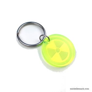 May include: A neon green keychain with a yellow radiation symbol. The keychain is round and attached to a silver metal key ring. The image is set against a white background.
