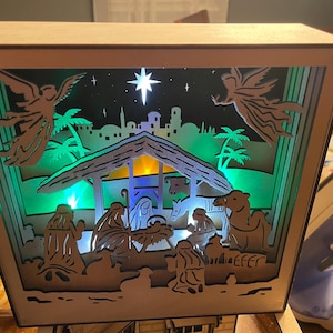 May include: A wooden nativity scene with a layered design, featuring silhouettes of angels, a star, and the holy family. The scene is illuminated with green and yellow lights, creating a warm glow. The overall theme is religious and festive.