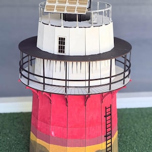 May include: A miniature lighthouse model featuring a red base with a yellow and brown bottom section, a white midsection with a black railing, and a top level with a black roof and solar panels. A black ladder is attached to the side of the red base.